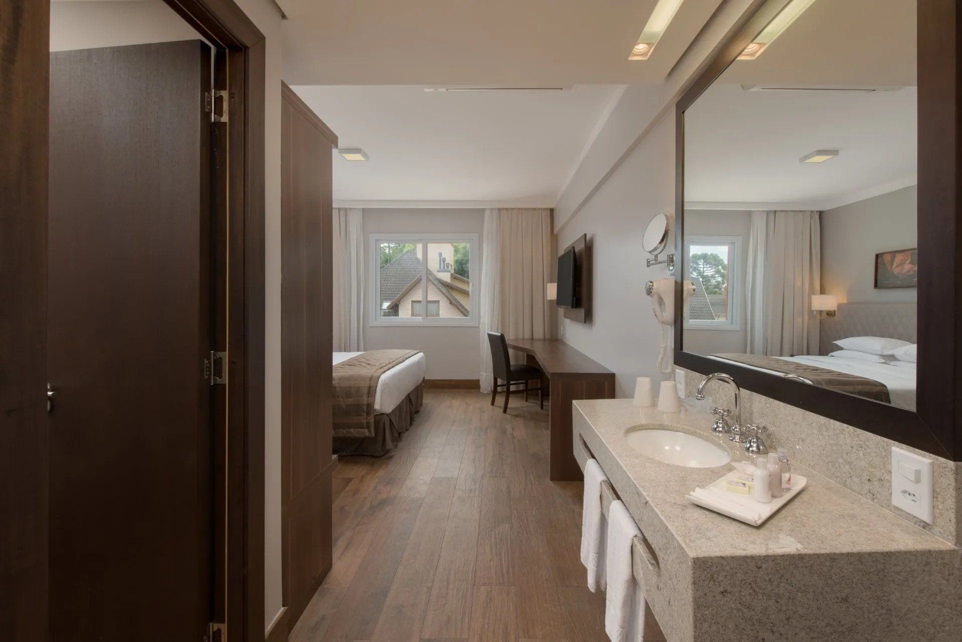 Quarto de hotel moderno com duas camas de solteiro, escrivaninha, lavatório no banheiro e janela ampla com vista para a cidade.