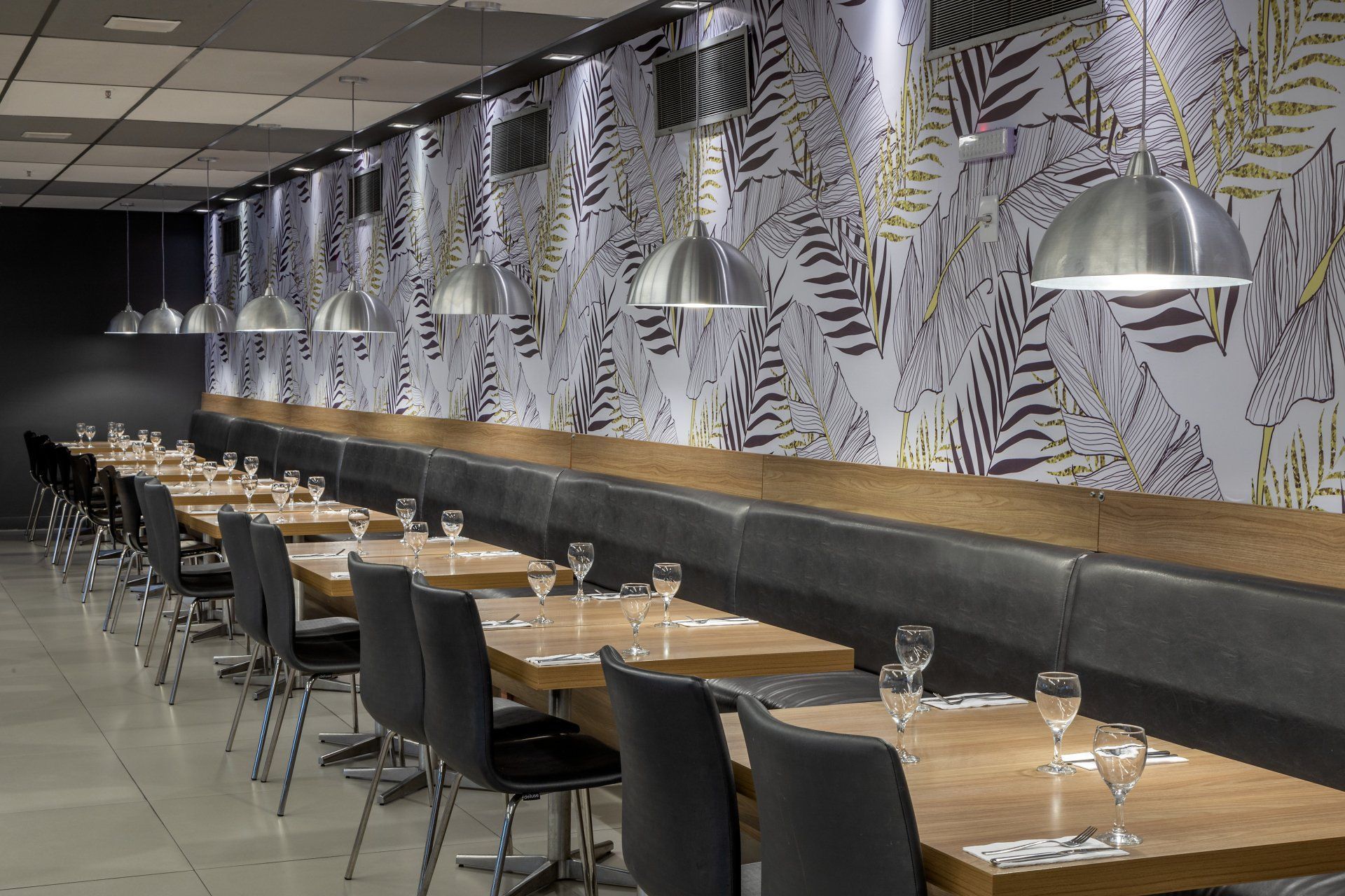 Área de jantar vazia de um restaurante moderno com parede estampada, mesas de madeira, cadeiras pretas e luminárias pendentes no teto.