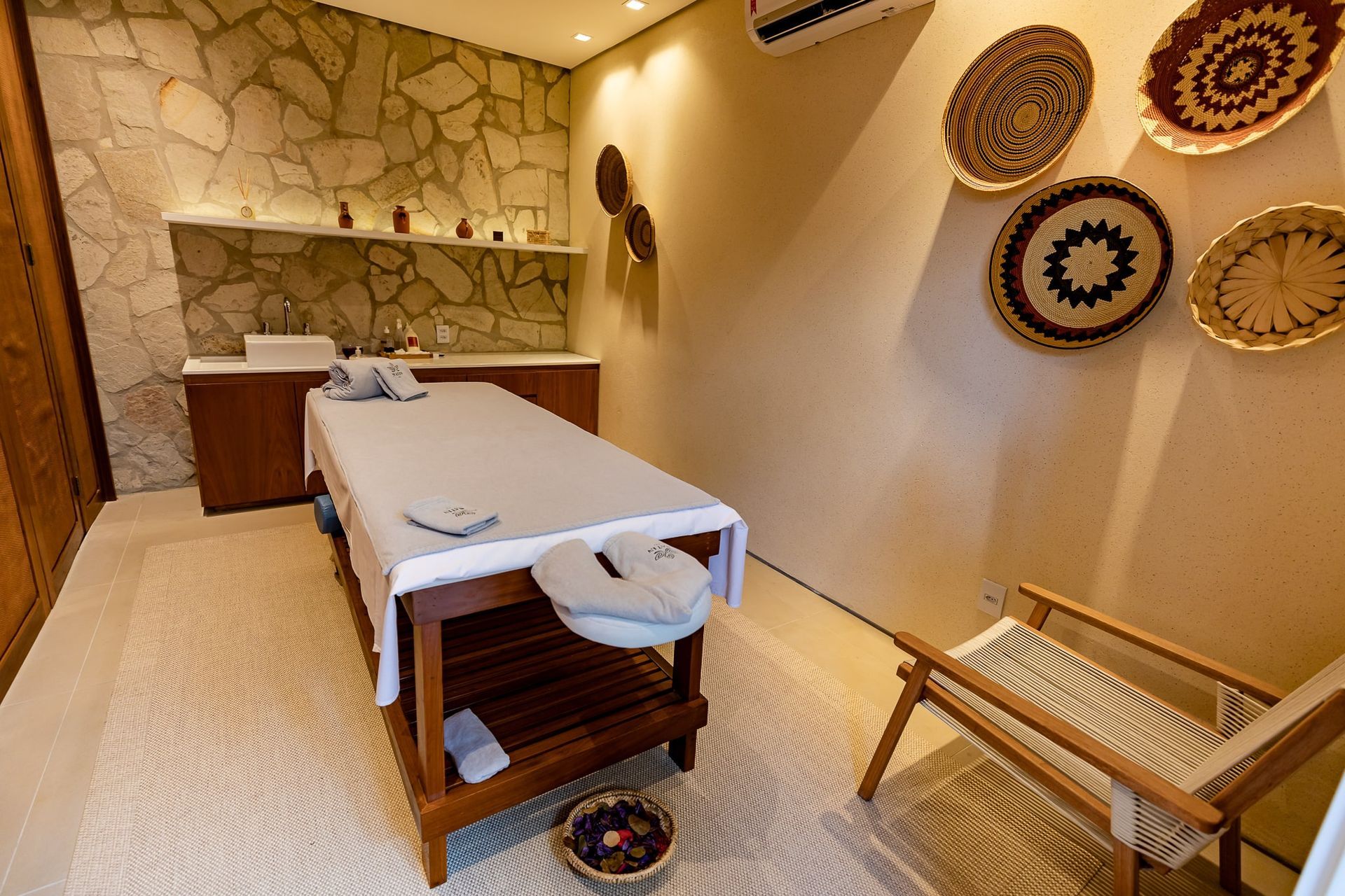 Sala de massagem de spa com paredes em tons creme, maca, cadeira de vime e cestos decorativos na parede.