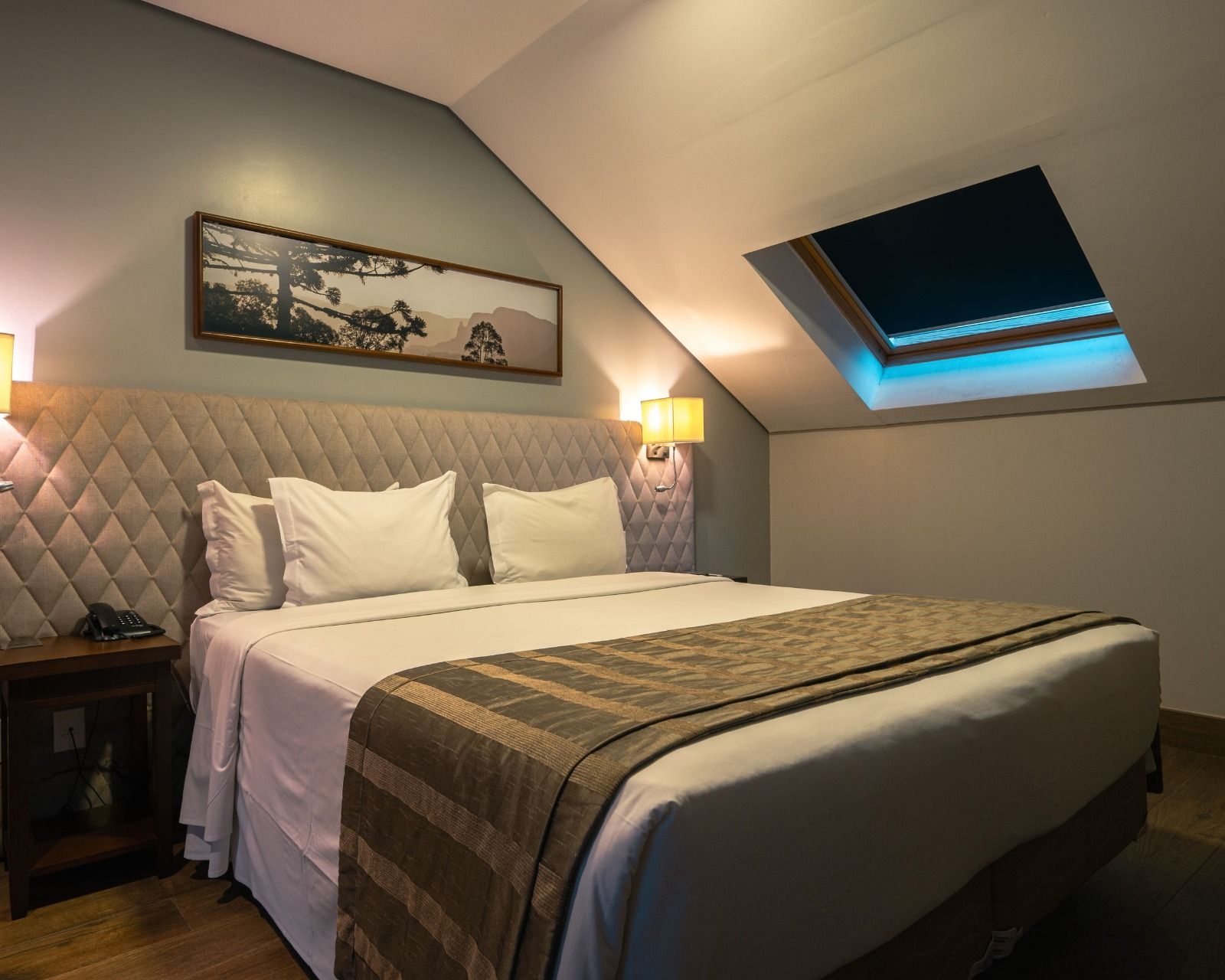 Quarto aconchegante no sótão com cama king size, cabeceira estofada, abajures e claraboia no teto.