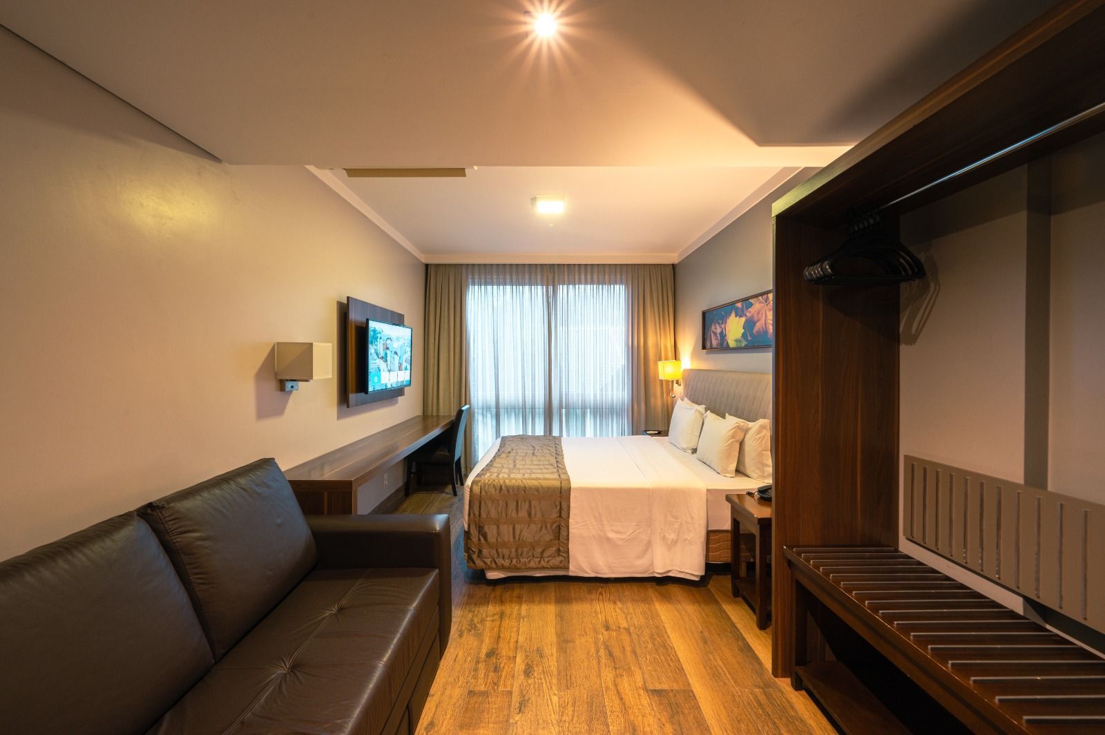 Quarto de hotel moderno com cama, sofá, TV, escrivaninha e cortinas amplas na janela.