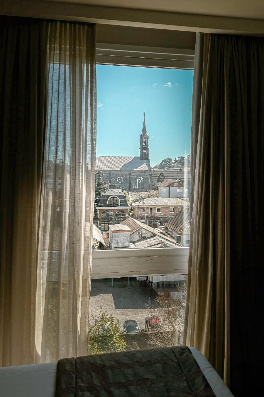 Vista através de uma janela com cortinas para uma paisagem urbana iluminada pelo sol e uma torre de igreja ao longe sob um céu azul.