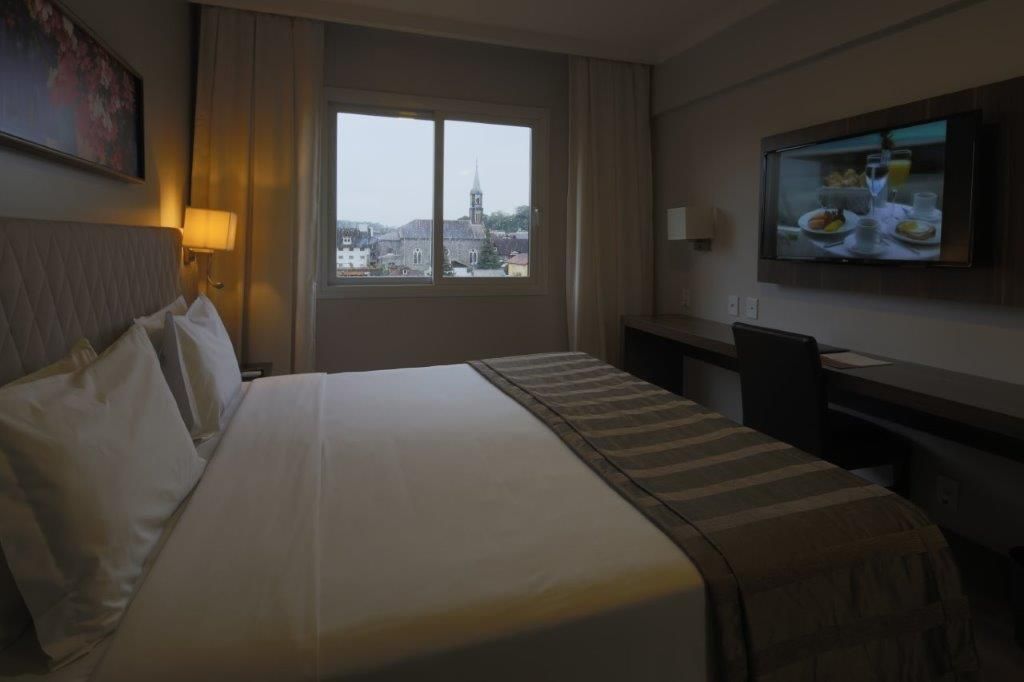 Quarto de hotel com cama arrumada, vista da janela e TV na parede.
