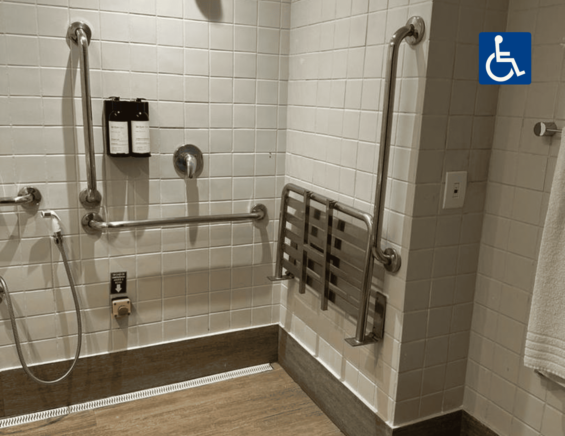 Chuveiro acessível com azulejos, barras de apoio, ducha manual, assento dobrável e placa com o símbolo de cadeira de rodas.