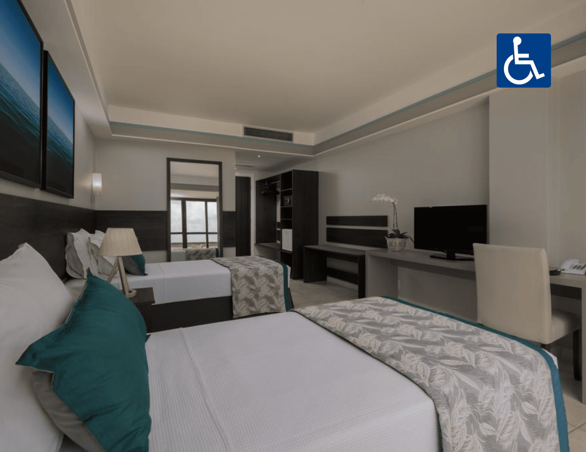 Quarto de hotel acessível com duas camas, escrivaninha, TV e placa de acesso para cadeirantes na parede.