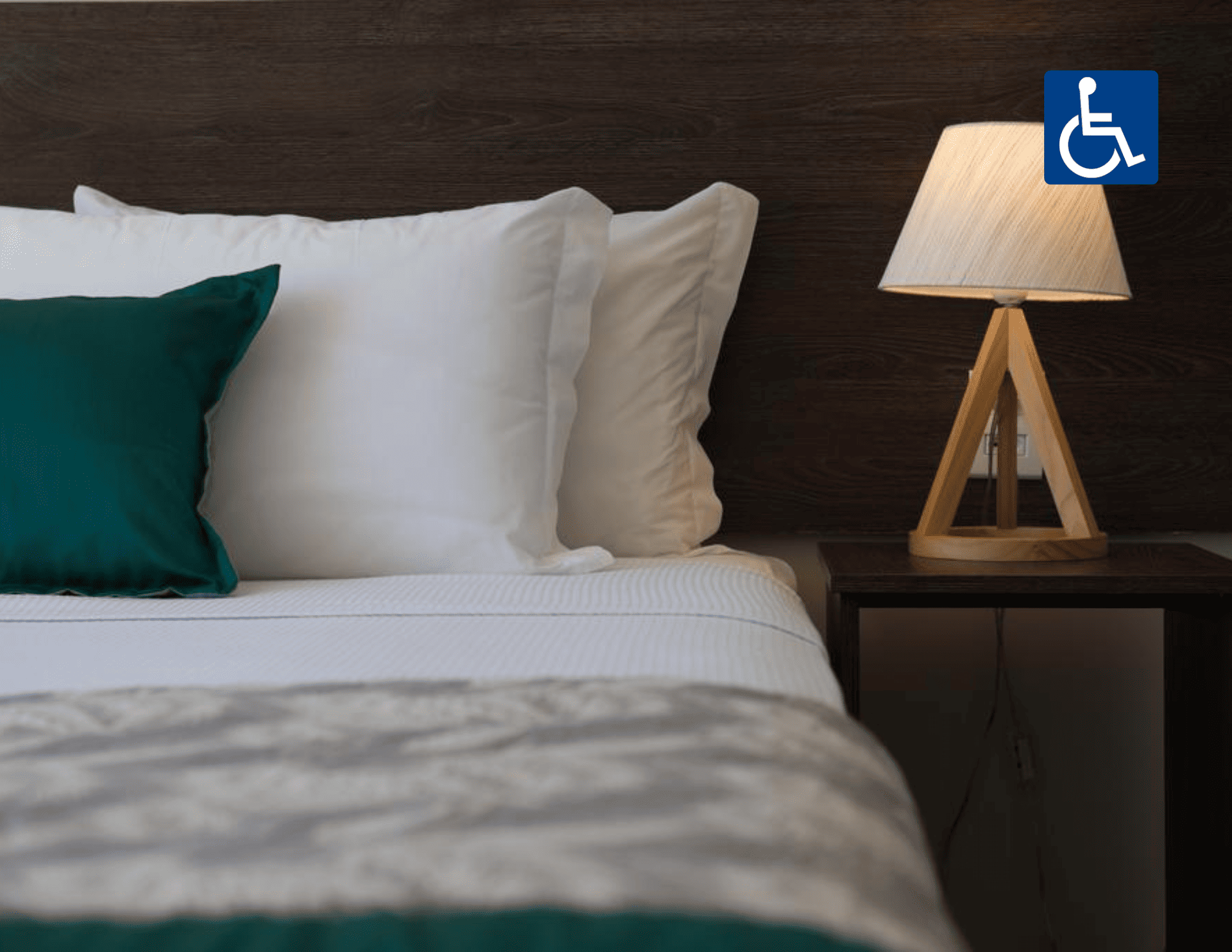 Quarto com cama arrumada, travesseiro azul-petróleo e abajur aceso sobre a mesa de cabeceira.