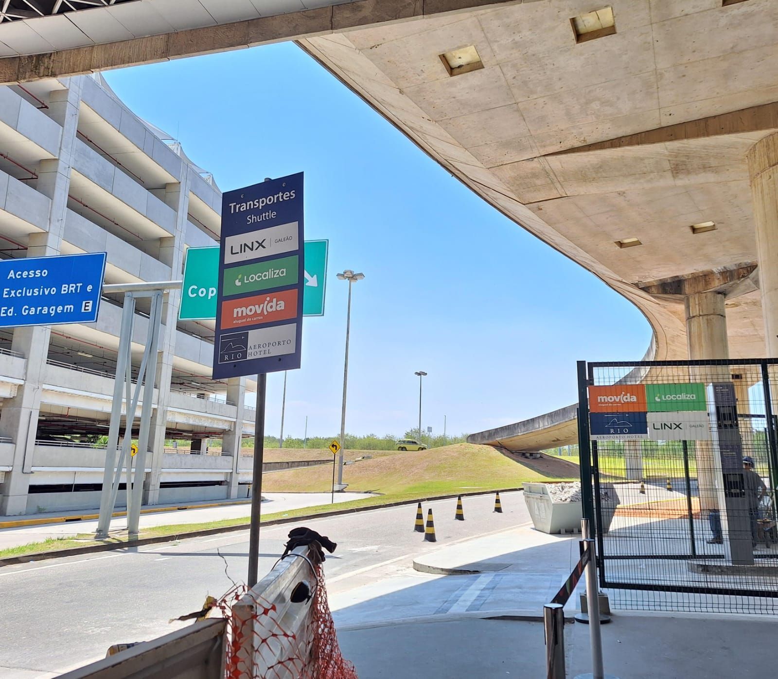 Entrada do aeroporto junto ao meio-fio com placas indicativas, cobertura de concreto e céu azul.