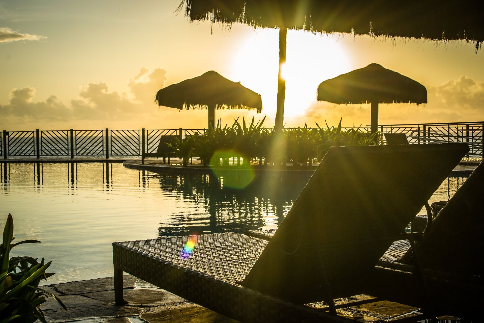 Pôr do sol à beira da piscina com espreguiçadeiras, guarda-sóis de palha e água calma refletindo a luz dourada.