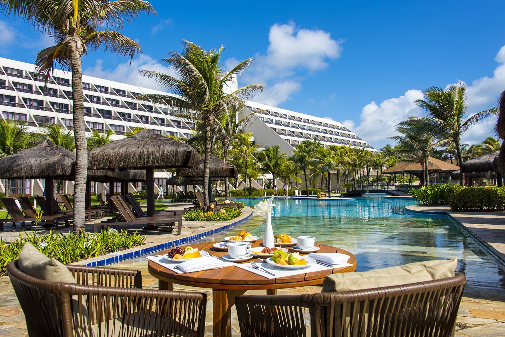 Tropical resort pool with palm trees, blue sky, and a breakfast tray on a wicker table by the water.