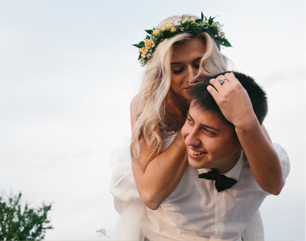 Casal sorridente se abraçando ao ar livre, a mulher com uma coroa de flores apoiada nos ombros do homem.