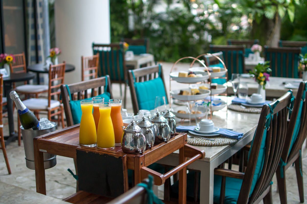 Conjunto de mesa e cadeiras para área externa, estilo café, com cadeiras verde-azuladas, louça branca, bules de chá prateados e jarras de suco amarelas.