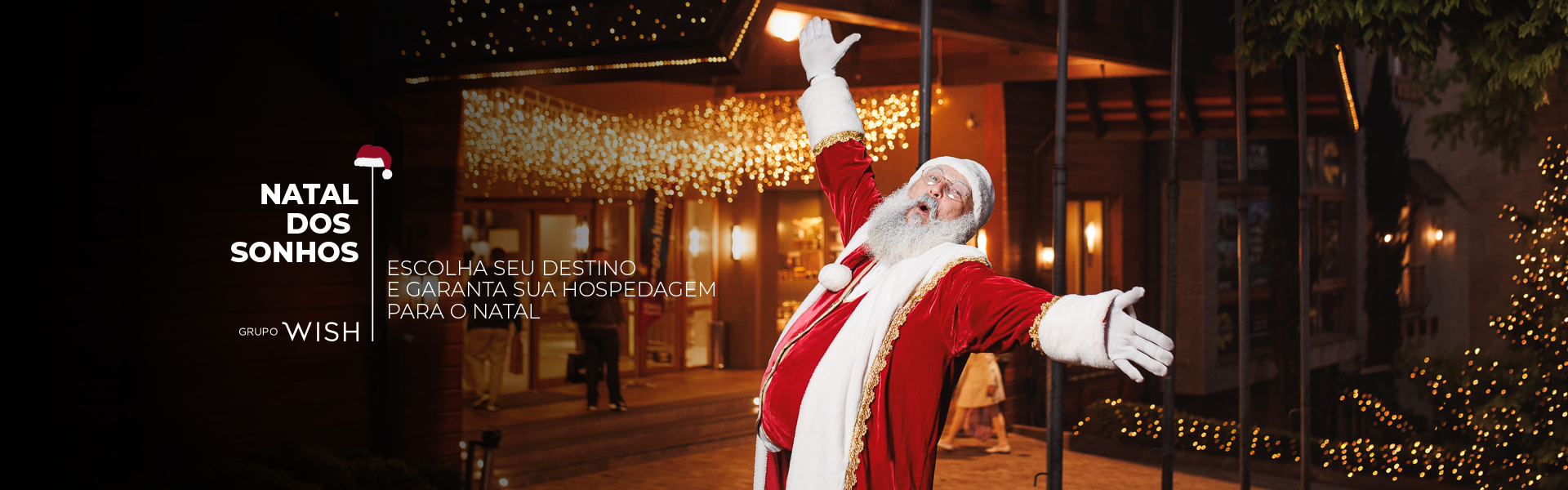 Papai Noel de casaco vermelho cumprimenta a todos em frente a um prédio iluminado à noite, com a mensagem 
