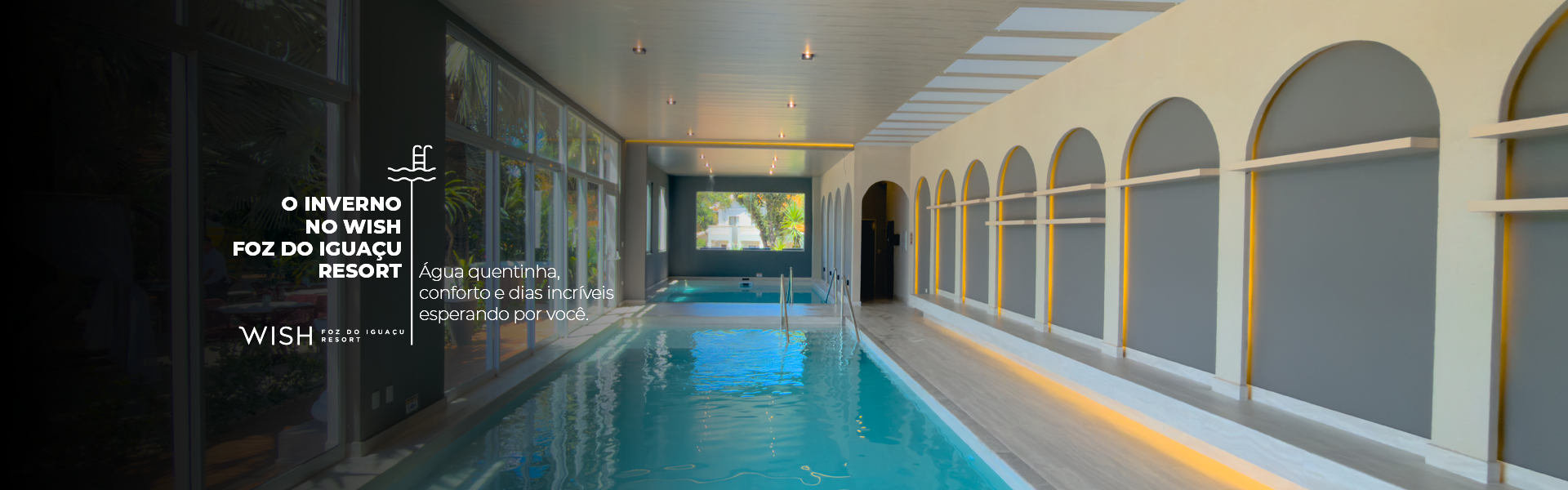 Piscina coberta com água azul, janelas em arco e um caminho de azulejos ao lado.