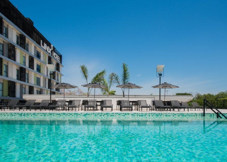 Piscina moderna de hotel com água turquesa, espreguiçadeiras e céu azul ao lado de um prédio de vários andares.