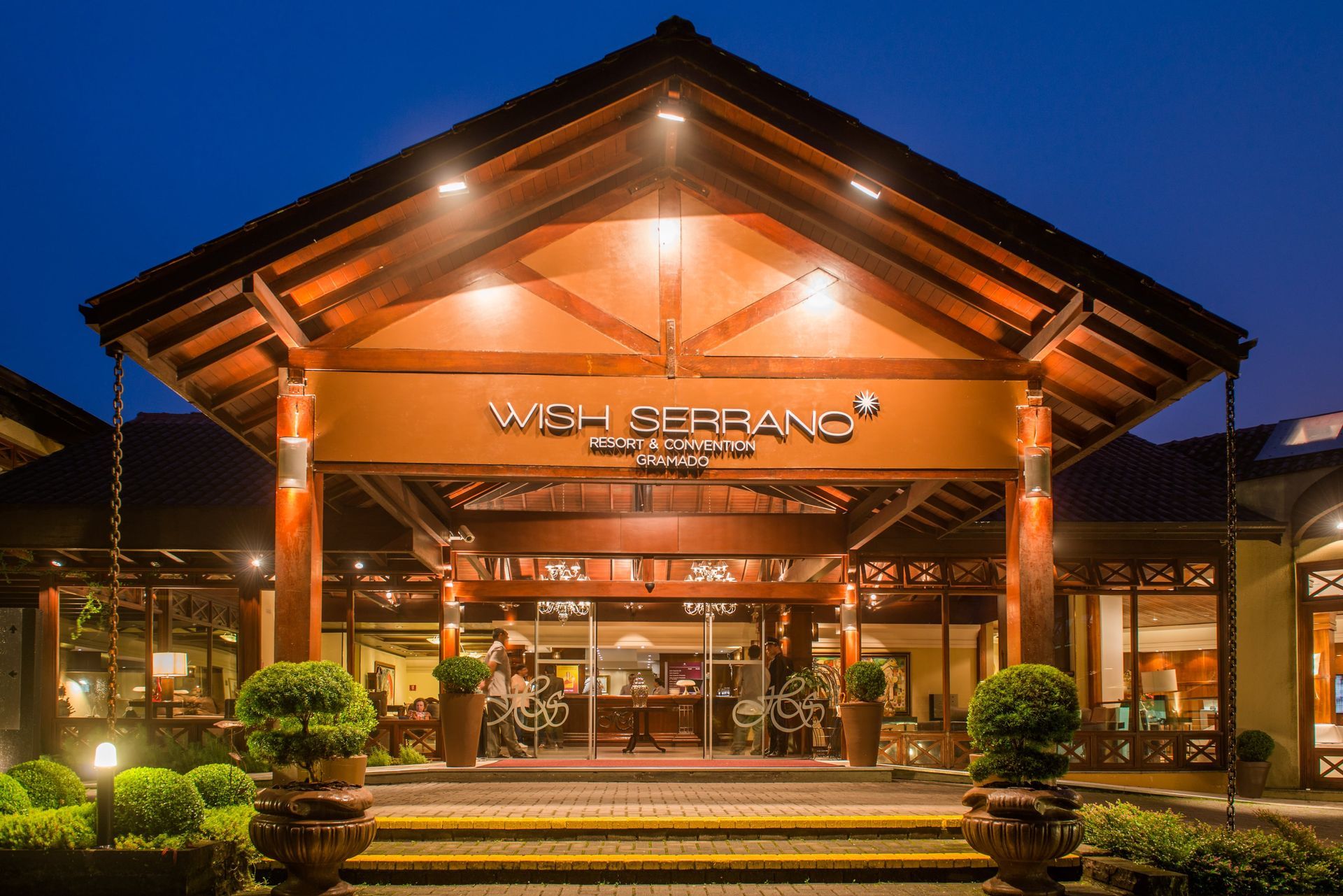 Entrada do resort bem iluminada à noite, com teto de madeira, portas de vidro e luzes laranja aconchegantes.
