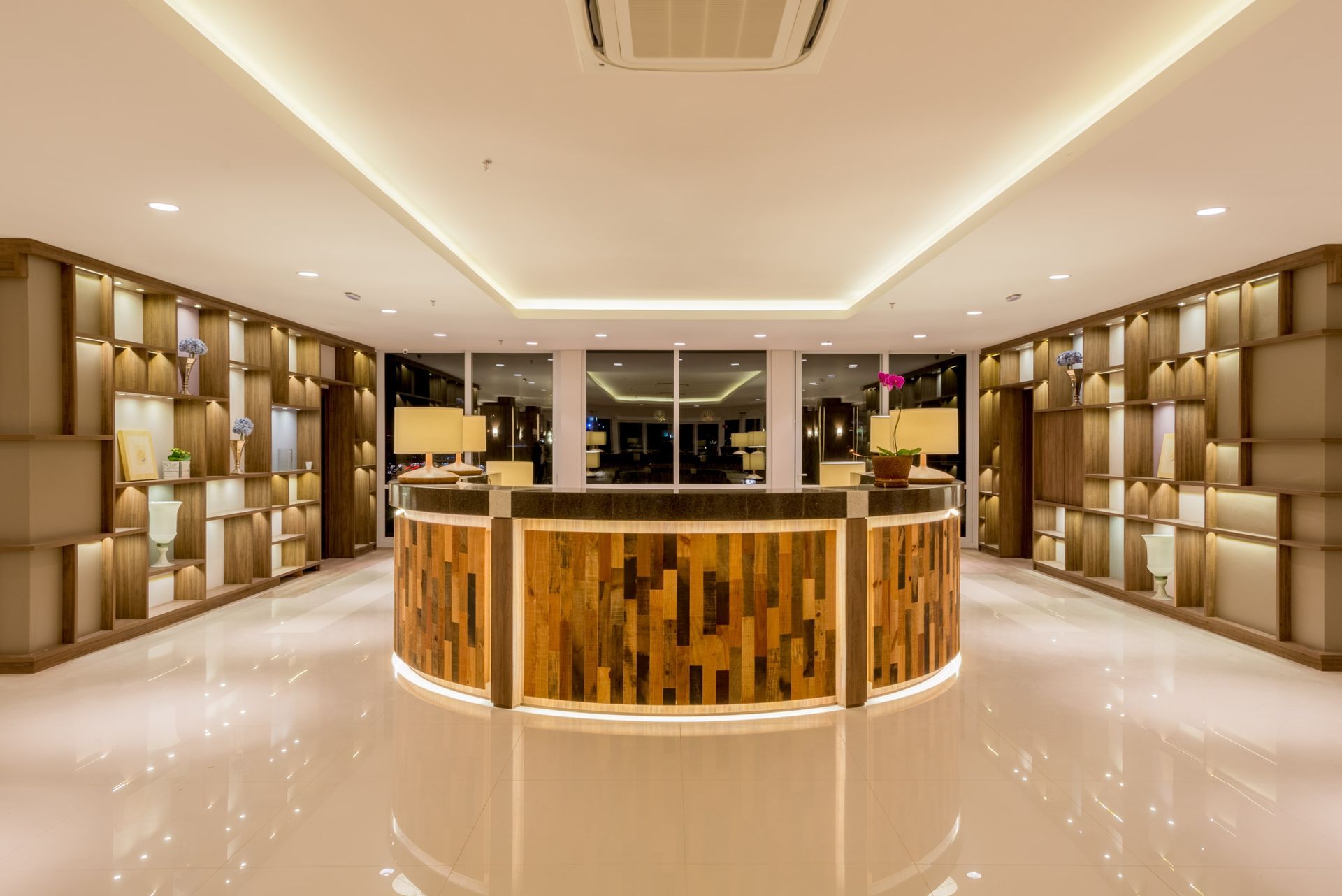 Hall de entrada moderno do hotel com balcão de recepção redondo de madeira, prateleiras douradas e iluminação embutida aconchegante.