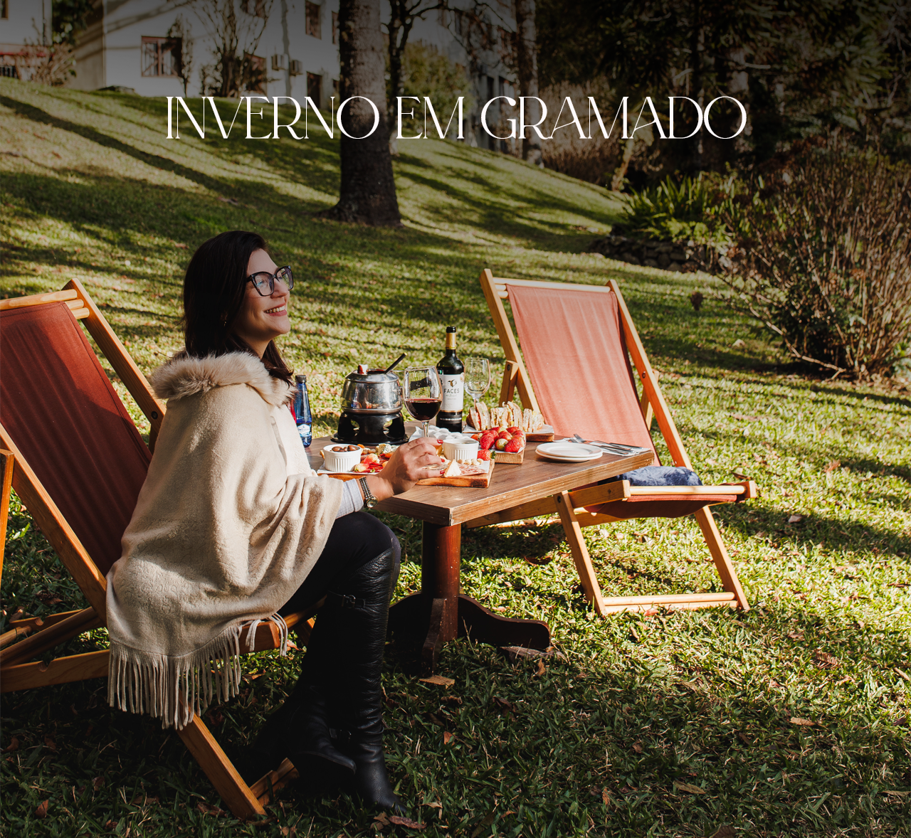 Uma mulher com um poncho creme senta-se a uma mesa ao ar livre com vinho e petiscos num jardim ensolarado.