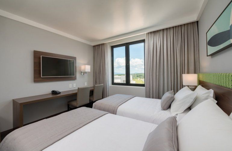 Quarto de hotel moderno com duas camas, escrivaninha, TV de parede e janela com cortinas.