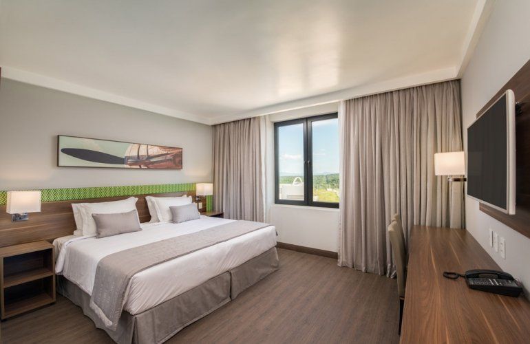 Quarto de hotel moderno com cama, escrivaninha, TV de parede, janela e cortinas.