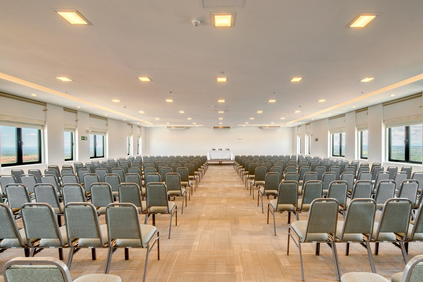 Sala de conferências vazia com fileiras de cadeiras cinzentas, piso de madeira e luzes embutidas brilhantes no teto.