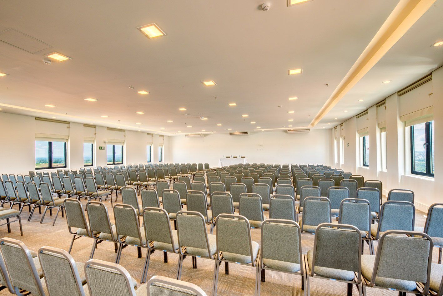 Sala de conferências vazia com fileiras de cadeiras cinzentas voltadas para a frente e janelas amplas ao longo da parede.
