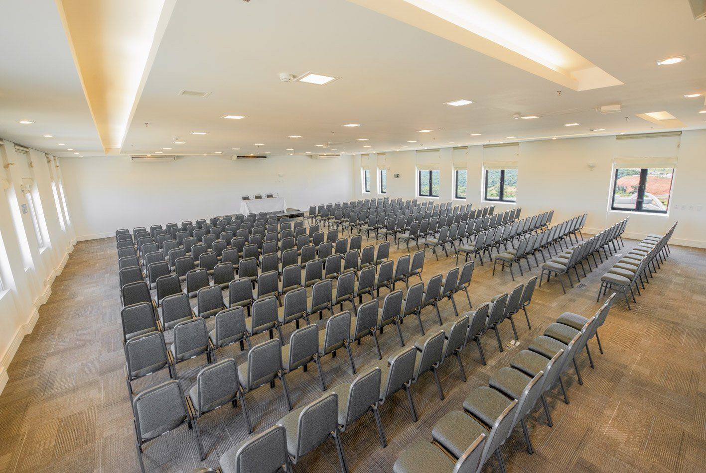 Sala de aula moderna e vazia, com fileiras de cadeiras cinzentas e paredes brancas, iluminada por luzes de teto e janelas.