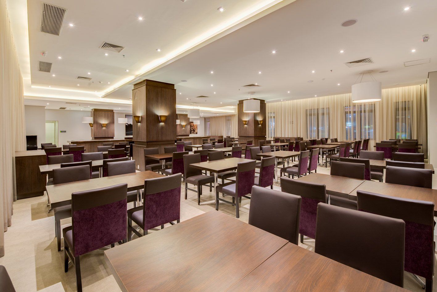 Sala de jantar vazia de um restaurante moderno com cadeiras roxas, mesas de madeira e iluminação aconchegante no teto.