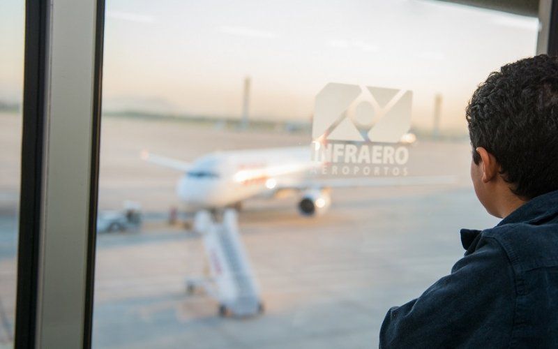 Pessoa observando um avião de passageiros na pista do aeroporto através de uma janela.