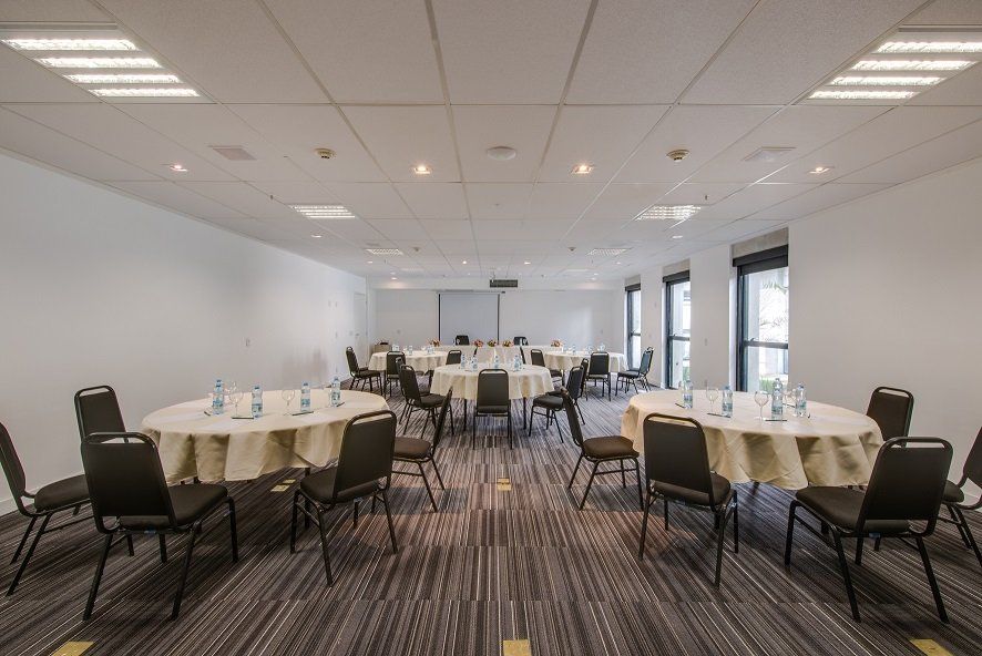 Sala de conferências com mesas redondas, cadeiras pretas e um quadro branco na frente.