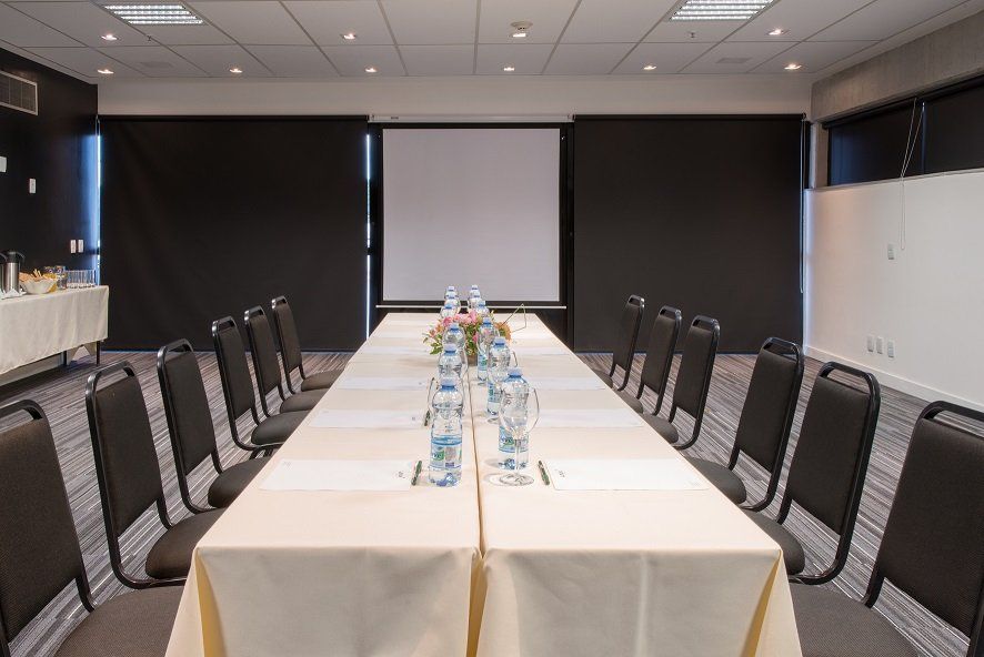 Sala de conferências com mesa comprida, garrafas de água, cadeiras pretas e uma tela de projeção ao fundo.