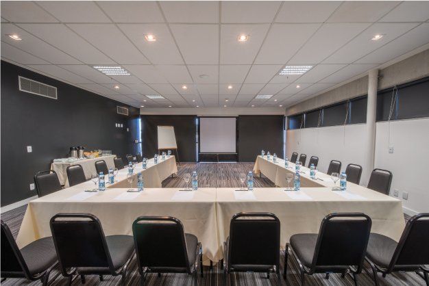 Sala de conferências com mesa em formato de U, cadeiras pretas, garrafas de água e tela de projeção.