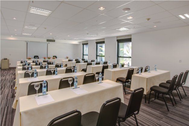 Sala de conferências com fileiras de mesas e cadeiras, cartazes de papel em cada assento e grandes janelas que permitem a entrada de luz natural.