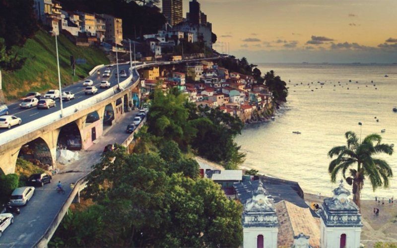 Rua costeira ao pôr do sol, com carros em uma estrada na encosta ao lado de uma orla ladeada por palmeiras.