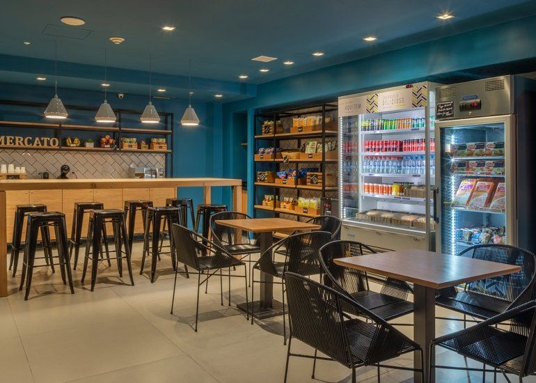 Interior de café azul com mesas altas, cadeiras pretas, luminárias pendentes e vitrine refrigerada.