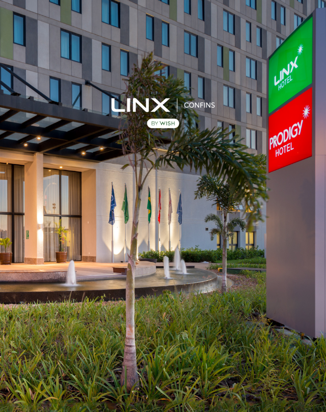 Entrada do hotel Linx com fonte, palmeiras e sinalização iluminada em verde e vermelho ao entardecer.