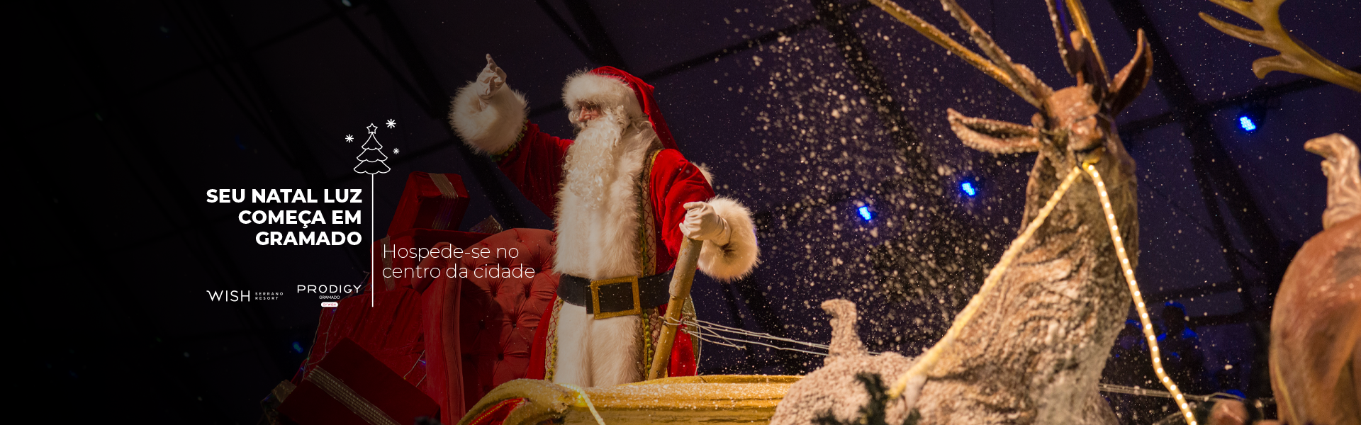 Papai Noel de roupa vermelha acena ao lado de uma rena iluminada e decorações em forma de estrela em um palco festivo.