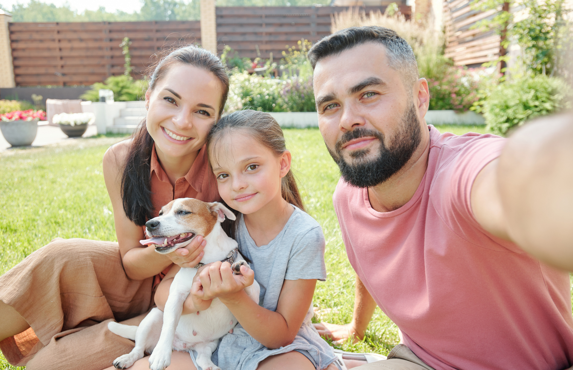 Família tirando uma selfie ao ar livre, sorrindo com um cachorrinho em um quintal ensolarado.