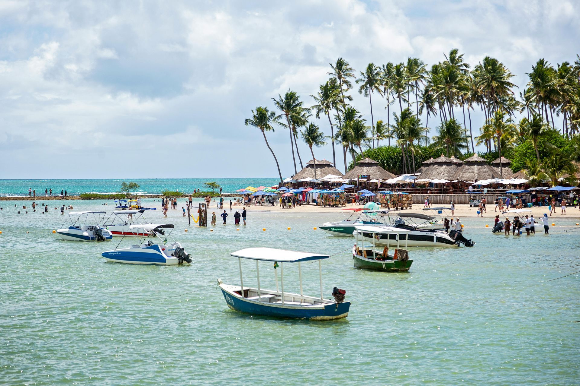 Tropical beach with anchored boats, swimmers, and palm trees under a cloudy sky