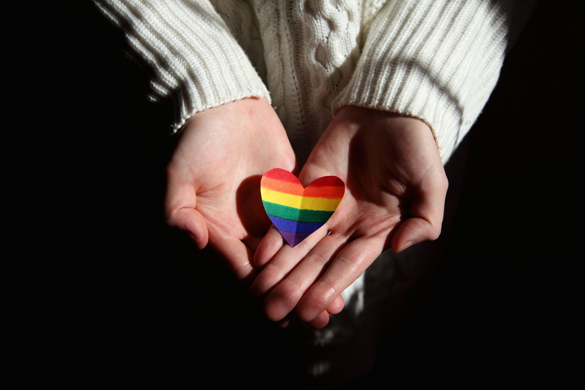 Hands holding a small rainbow heart against a dark background