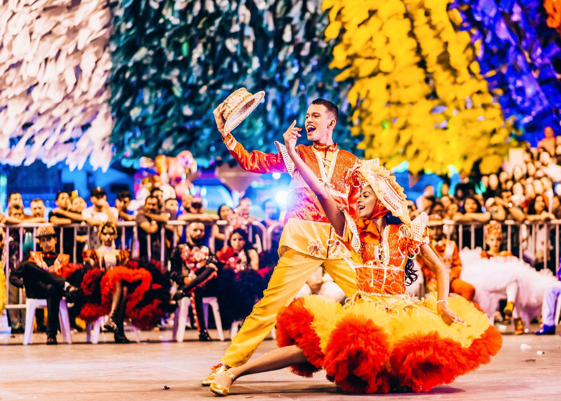 Dancers in colorful costumes perform on stage before a seated audience, with bright lights and a vivid backdrop.