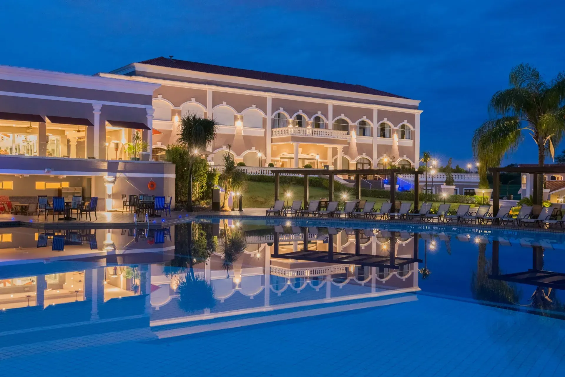 Resort de luxo com piscina iluminada e imponente edifício branco refletido ao entardecer.