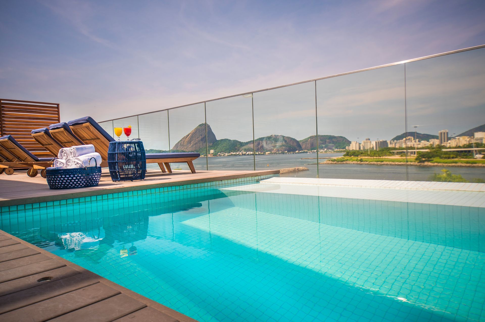 Piscina infinita na cobertura com espreguiçadeiras, com vista para as montanhas e para o porto ao pôr do sol.