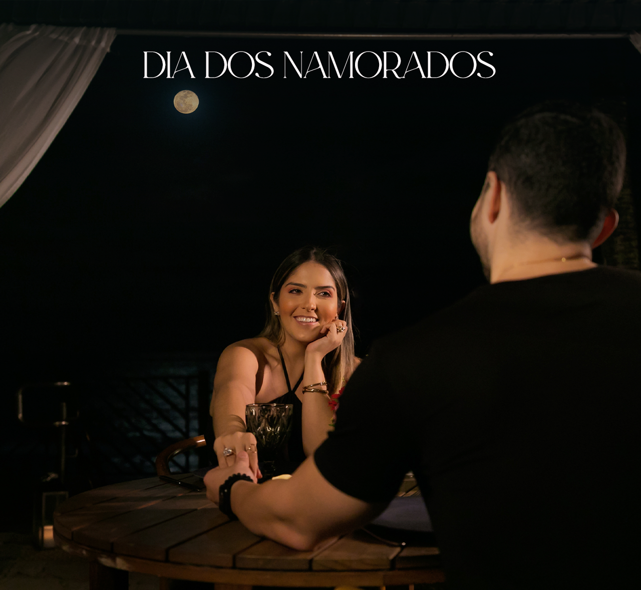 Casal sentado em uma mesa romântica com iluminação suave sob uma placa de “Dia dos Namorados”.