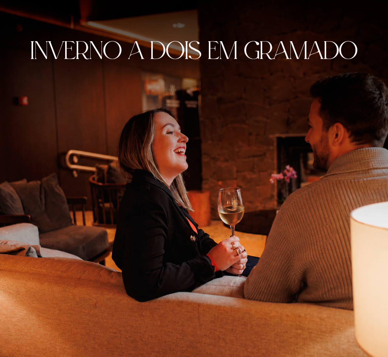 Casal conversando em um sofá perto da lareira em uma cabana aconchegante de inverno, com a inscrição “INVERNO A DOIS EM GRAMADO” acima.
