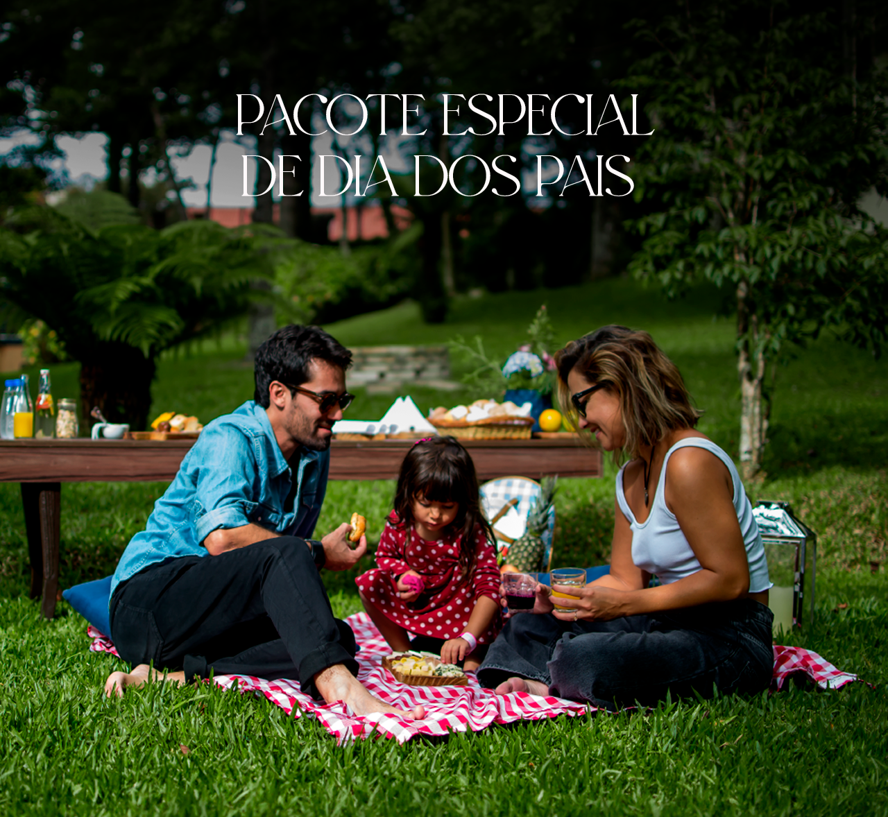 Family picnic on a pink blanket in a park with a “Pacote especial de Dia dos Pais” sign.