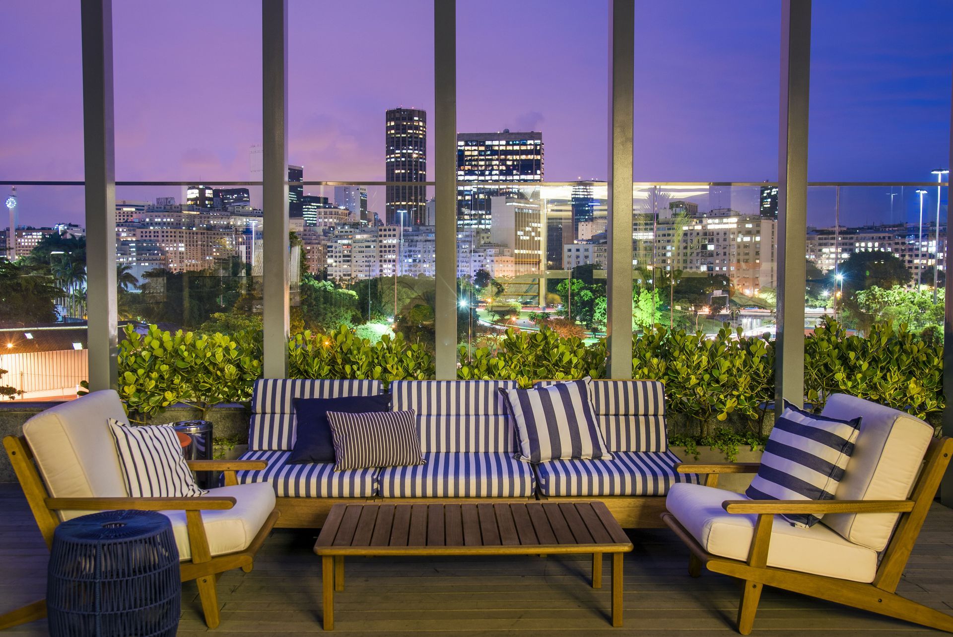 Lounge ao ar livre com terraço, assentos listrados e vista para o horizonte da cidade ao entardecer.