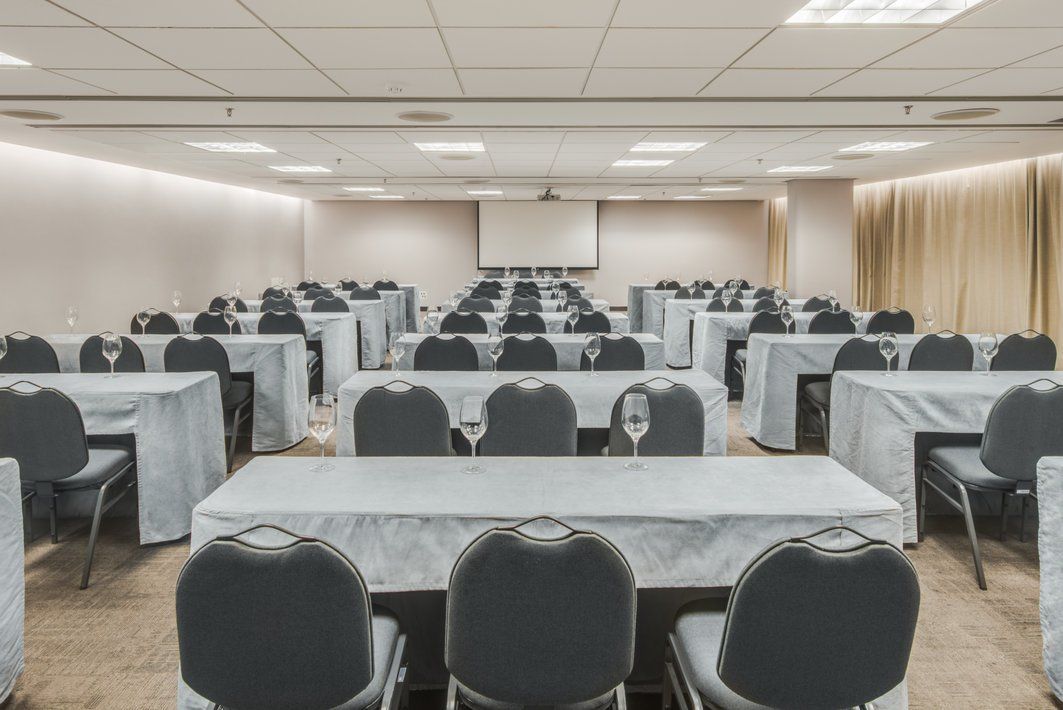 Sala de conferências vazia com fileiras de mesas e cadeiras pretas sob luzes fortes no teto.