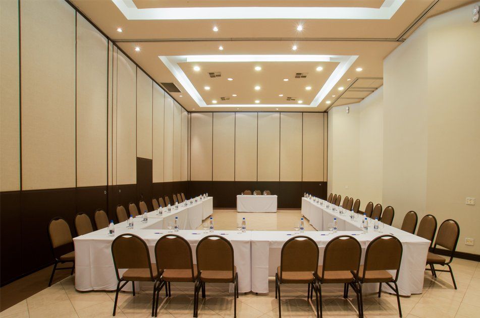 Sala de conferências vazia com mesas em formato de U, toalhas de mesa brancas, cadeiras marrons e iluminação embutida no teto.