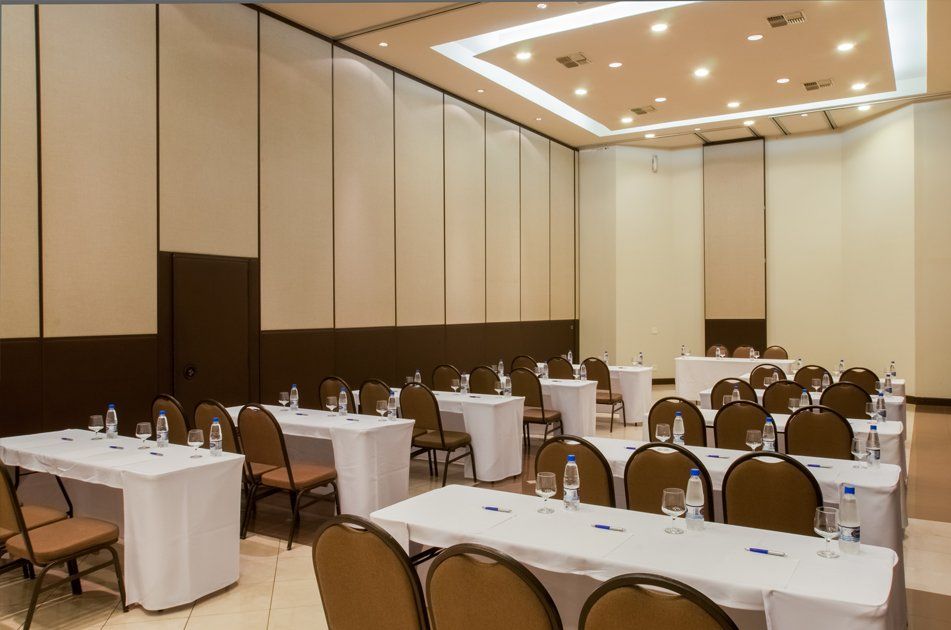 Sala de conferências com fileiras de mesas cobertas com toalhas brancas e cadeiras marrons sob luzes de teto aconchegantes.