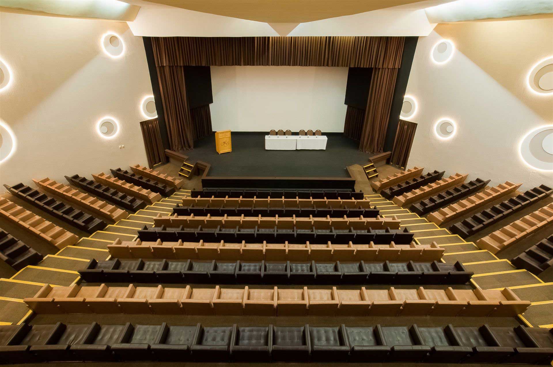 Auditório vazio com fileiras de assentos voltadas para um palco e tela de projeção, iluminado por apliques de parede.